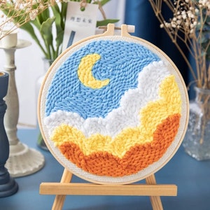 Peut inclure: Un cercle de broderie rond avec un ciel bleu, des nuages blancs et des montagnes orange et jaunes. Une lune en croissant jaune est dans le ciel. Le cercle est sur un chevalet en bois.