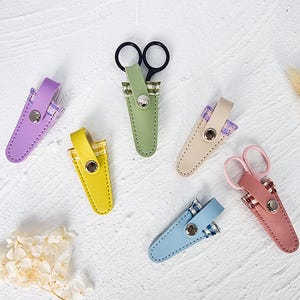 May include: A collection of small scissors with black handles and colourful leather sheaths. The sheaths are in various colours, including purple, yellow, green, beige, blue, and pink. Each sheath has a snap closure.