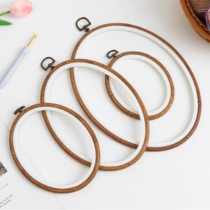 May include: Set of three oval embroidery hoops with wooden frames and white inner rings. The hoops are nested inside each other, with the largest hoop at the bottom and the smallest hoop at the top.