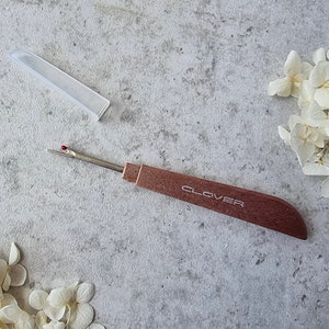 May include: A small, wooden-handled thread snipper with a red tip and a silver blade. The snipper is labelled "CLOVER" and is sitting on a grey surface.