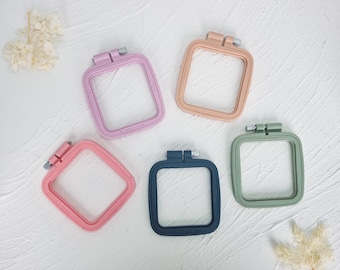 May include: Five square embroidery hoops in different colours: pink, light pink, peach, dark blue, and green. The hoops are all the same size and have a metal screw closure.