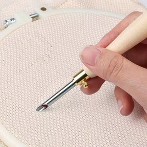 May include: A close-up of a wooden embroidery needle threader with a gold metal tip and a red string attached to the end. The needle threader is being held over a white embroidery hoop.