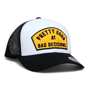 May include: A white and black trucker hat with a curved brim. The hat features a yellow patch with the text "PRETTY GOOD at BAD DECISIONS" in black lettering. The back of the hat is black mesh.