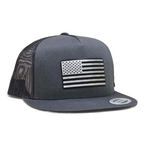 May include: A dark grey trucker cap with a black mesh back and a flat peak. The front panel displays a silver and white American flag patch. The cap has a snapback closure for an adjustable fit.