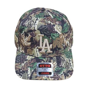 May include: A camouflage baseball cap with the letters "LA" embroidered on the front. The cap features a classic fit and is made of premium fabric. The cap has a curved brim and a button on top.