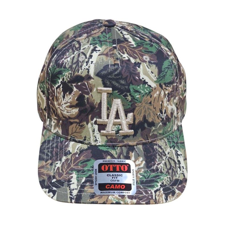 May include: A camouflage baseball cap with the letters "LA" embroidered on the front. The cap features a classic fit and is made of premium fabric. The cap has a curved brim and a button on top.