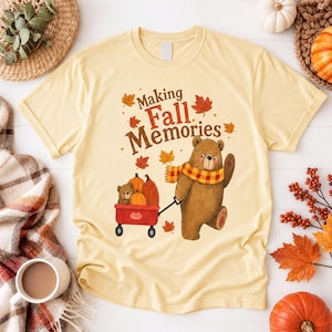 Może przedstawiać: Jasnożółty t-shirt z grafiką przedstawiającą brązowego niedźwiedzia w żółto-czerwony szalik w kratę, ciągnącego czerwony wózek wypełniony dyniami i małym niedźwiadkiem. Powyżej znajduje się brązowy napis "Making Fall Memories".