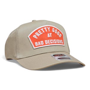 May include: Tan trucker hat with a mesh back and a curved brim. The front patch is orange with white text that reads "PRETTY GOOD AT BAD DECISIONS."