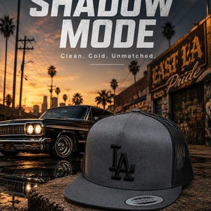 May include: A grey and black trucker hat featuring the letters LA in black embroidery on the front panel. The hat has a flat brim with a circular sticker and a mesh back. The text SHADOW MODE, Clean. Cold. Unmatched. and AVAILABLE NOW is visible.
