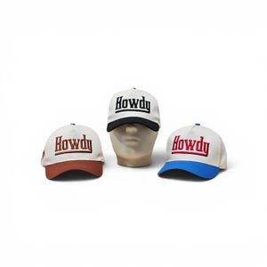 May include: Three baseball caps with off-white crowns and embroidered text reading Howdy. The left cap has a brown brim, the centre cap on a mannequin head has a black brim, and the right cap has a blue brim. Each features structured panels.