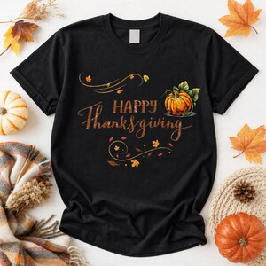 May include: A black short-sleeved t-shirt featuring the text Happy Thanksgiving in a cursive font. The design includes a graphic of an orange pumpkin with green leaves and decorative swirling lines accented with small autumn-coloured leaves.