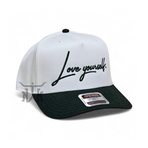 Puede incluir: Gorra de béisbol blanca y negra con la frase "Love yourself" bordada en escritura negra. La gorra tiene una visera negra y un botón negro en la parte superior. Ajuste clásico.