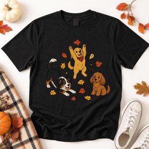 Può includere: Una t-shirt nera a maniche corte con una stampa grafica di tre cani che giocano tra le foglie autunnali che cadono. L'illustrazione include un cane bianco e nero, un cane dorato con una bandana rossa e un cane marrone con una bandana gialla.