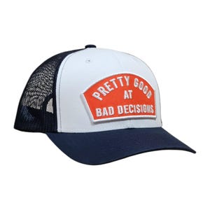 May include: A white and navy blue trucker hat with a mesh back. The hat features a patch with the words "PRETTY GOOD AT BAD DECISIONS" in white lettering on an orange background.