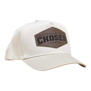 May include: A cream-coloured baseball cap with a curved peak. The cap has a brown leather patch with the word "CHOSEN" and the text "1 PETER 2:9" in a dark font. The cap is made of a durable fabric.