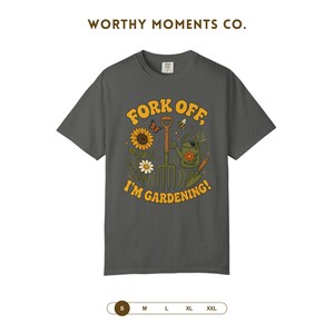 Può includere: Una t-shirt a maniche corte grigio antracite con una stampa grafica recante il testo "FORK OFF, I'M GARDENING!" circondato da illustrazioni di un girasole, una margherita, una forca da giardino, un annaffiatoio, una paletta, una farfalla e un'ape.