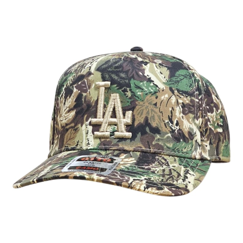 May include: A camouflage baseball cap with the letters "LA" embroidered on the front. The cap has a curved peak and a button on top. The fabric has a pattern of green, brown, and tan, resembling a woodland design.