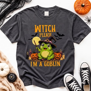 May include: A charcoal gray t-shirt featuring a graphic of a green frog wearing a witch hat, holding a wand, and surrounded by pumpkins, candles, and bats. The text reads WITCH PLEASE I'M A GOBLIN in yellow letters against a dark background.