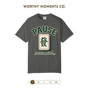 May include: A dark gray short-sleeve t-shirt featuring the text WORTHY MOMENTS CO. at the top. The center graphic displays the word PAUSE in green arched letters above a mahjong tile with the character Fa, followed by the text never mind...