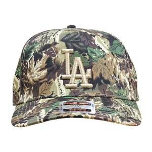 May include: A camouflage baseball cap with the letters "LA" embroidered on the front in a tan colour. The cap features a curved brim and a fabric pattern of green, brown, and tan leaves.