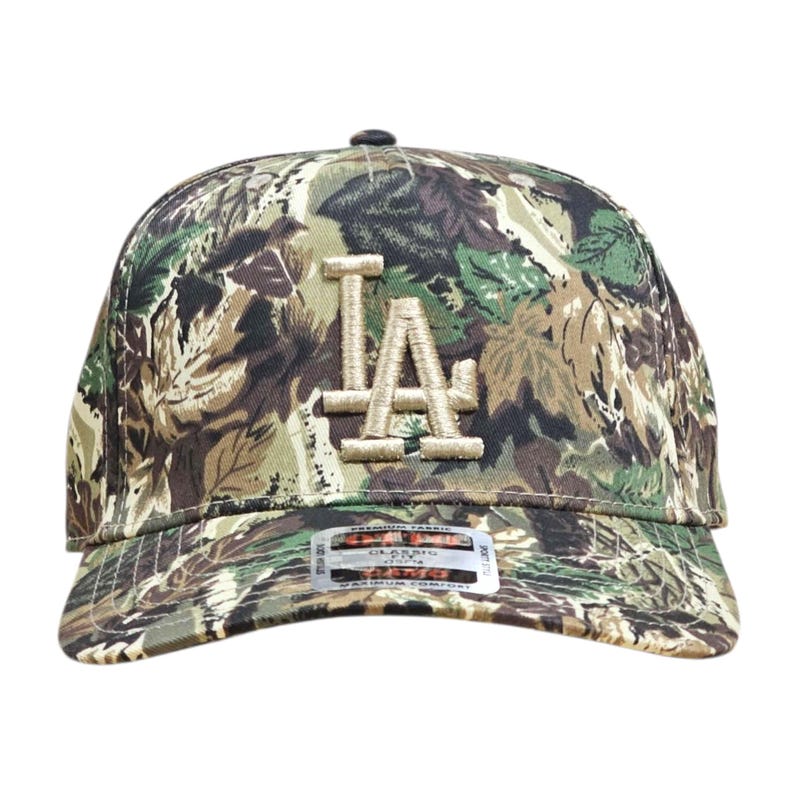 May include: A camouflage baseball cap with the letters "LA" embroidered on the front in a tan colour. The cap features a curved brim and a fabric pattern of green, brown, and tan leaves.