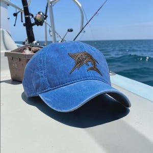 May include: A blue baseball cap featuring a marlin emblem. The cap is made of a soft fabric and has a curved peak. The marlin design is a detailed, brown patch on the front. The cap is on a boat, with fishing rods visible in the background.
