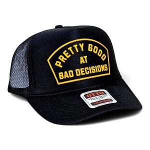 May include: Black trucker hat with a mesh back and a curved brim. The hat features a yellow and black patch that reads "PRETTY GOOD AT BAD DECISIONS". The hat has a white tag with black text.
