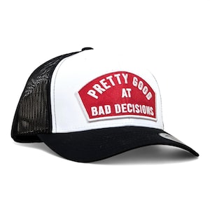 May include: A white and black trucker hat with a curved brim. The hat features a red patch with the words "PRETTY GOOD AT BAD DECISIONS" in white lettering. The hat has a black mesh back.