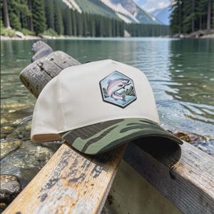Può includere: Cappellino da baseball color crema con visiera mimetica. Il cappellino presenta una toppa esagonale con un salmone che salta fuori dall'acqua. Lo sfondo mostra un lago e montagne.