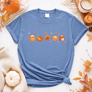 Peut inclure: Un t-shirt bleu à manches courtes avec une impression graphique centrale d'éléments sur le thème de l'automne, incluant une tasse avec de la crème fouettée, une citrouille, une feuille d'érable orange, une tartelette et une bougie allumée.