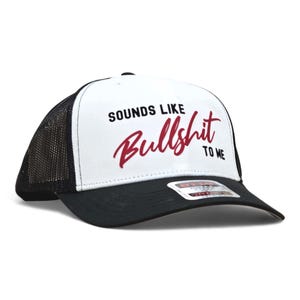 May include: A black and white trucker hat with the text "SOUNDS LIKE Bullshit TO ME" in black and red. The hat has a white front panel, a black brim, and a black mesh back.