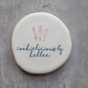May include: White round cookie with a blue and pink design of baking tools and the text "cookielicious by kellee".