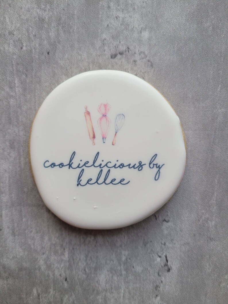 May include: White round cookie with a blue and pink design of baking tools and the text "cookielicious by kellee".