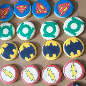 May include: A collection of 18 round sugar cookies decorated with various superhero logos. The logos include Superman, Batman, The Flash, Green Lantern, and more. The cookies are yellow, red, blue, green, and black.