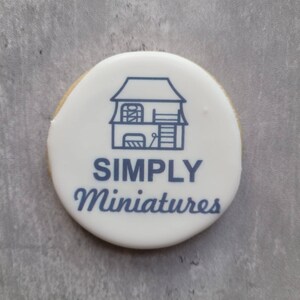 May include: A white round cookie with blue icing depicting a small house with a ladder and the text "SIMPLY Miniatures".
