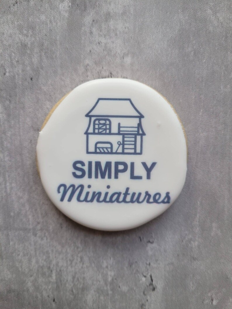 May include: A white round cookie with blue icing depicting a small house with a ladder and the text "SIMPLY Miniatures".