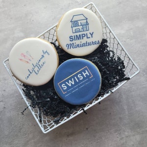May include: Three decorated sugar cookies in a white wire basket. The cookies are decorated with different designs. One cookie has a house with the text "SIMPLY Miniatures" on it. Another cookie has the text "cookies delicious by kellee" on it. The third cookie has the text "SWISH PHYSICAL THERAPY & PERFORMANCE" on it.