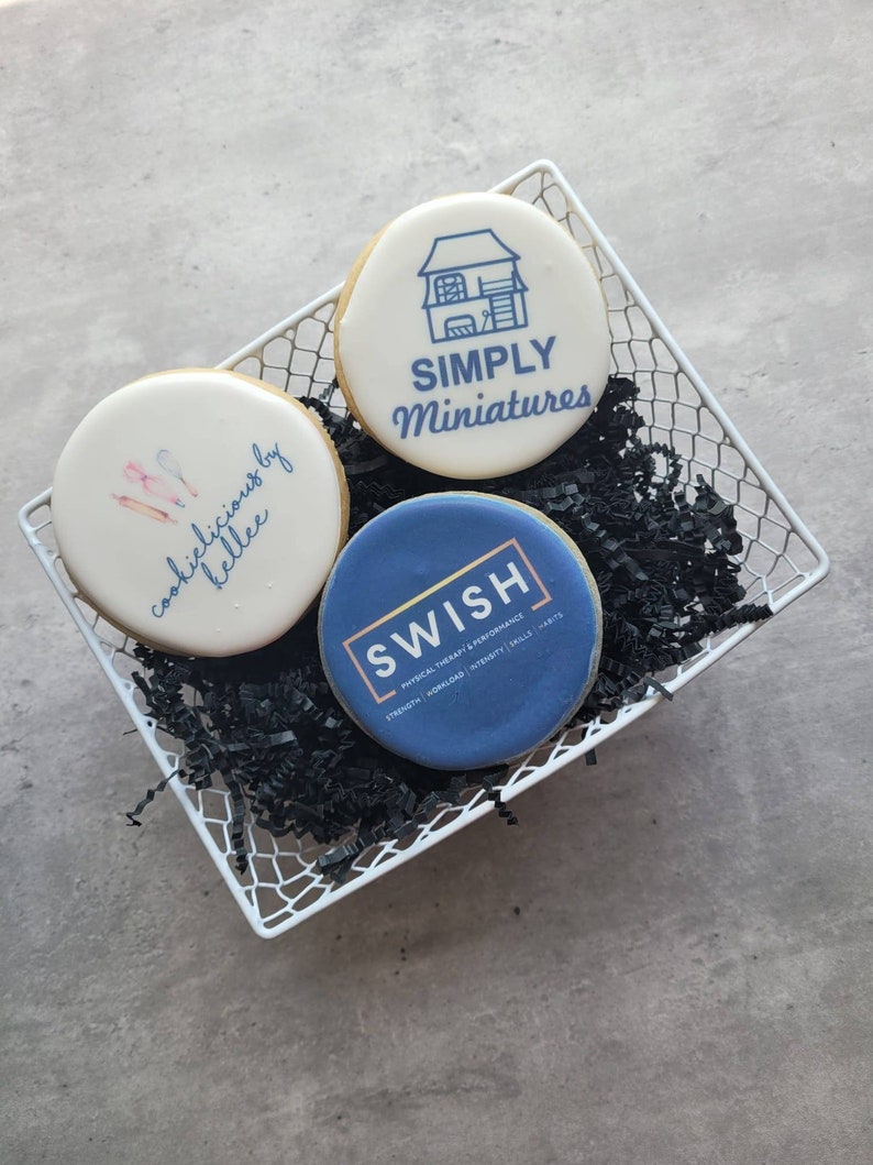 May include: Three decorated sugar cookies in a white wire basket. The cookies are decorated with different designs. One cookie has a house with the text "SIMPLY Miniatures" on it. Another cookie has the text "cookies delicious by kellee" on it. The third cookie has the text "SWISH PHYSICAL THERAPY & PERFORMANCE" on it.