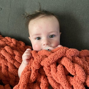 May include: A baby with brown hair is lying on a soft, orange chunky knit blanket. The baby is wearing a dummy and is looking directly at the camera.
