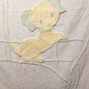 May include: A white crocheted baby blanket with a yellow puppy design. The puppy has blue eyes and a pink nose.