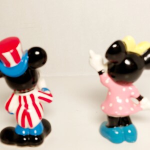 May include: A pair of small ceramic figurines of Mickey Mouse and Minnie Mouse. Mickey is wearing a red, white, and blue hat and a red, white, and blue striped shirt. Minnie is wearing a pink dress with white polka dots and a yellow bow.