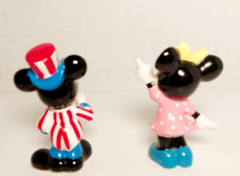 May include: A pair of small ceramic figurines of Mickey Mouse and Minnie Mouse. Mickey is wearing a red, white, and blue hat and a red, white, and blue striped shirt. Minnie is wearing a pink dress with white polka dots and a yellow bow.