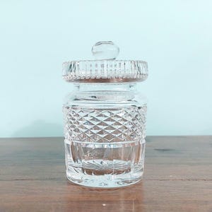May include: A clear, cut-glass jar with a lid. The jar has a diamond pattern on the body and a round knob on the lid. The jar is sitting on a wooden surface.