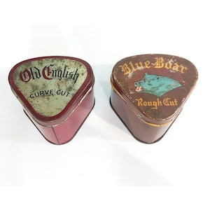 May include: Two triangular metal tobacco tins. The left tin is maroon with a weathered silver lid labeled Old English Curve Cut. The right tin is brown with a gold-toned lid featuring a teal boar illustration and the text Blue Boar Rough Cut.
