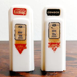 May include: Two white ceramic gas pump salt and pepper shakers. The left shaker has "CONOCO SUPER!" printed on the top and "CONOCO" printed on the bottom. The right shaker has "CONOCO" printed on the top.