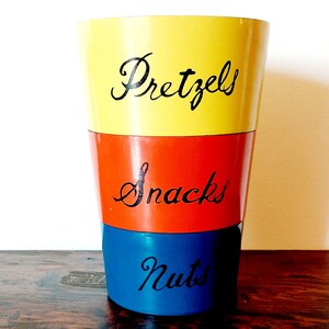 May include: A yellow, red, and blue vintage snack container with the words "Pretzels", "Snacks", and "Nuts" written in black.