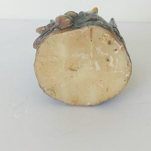 May include: The bottom of a ceramic figurine of a gnome sitting on a tree stump. The figurine is painted in shades of brown, green, and blue. The bottom of the figurine is white and has a rough texture.