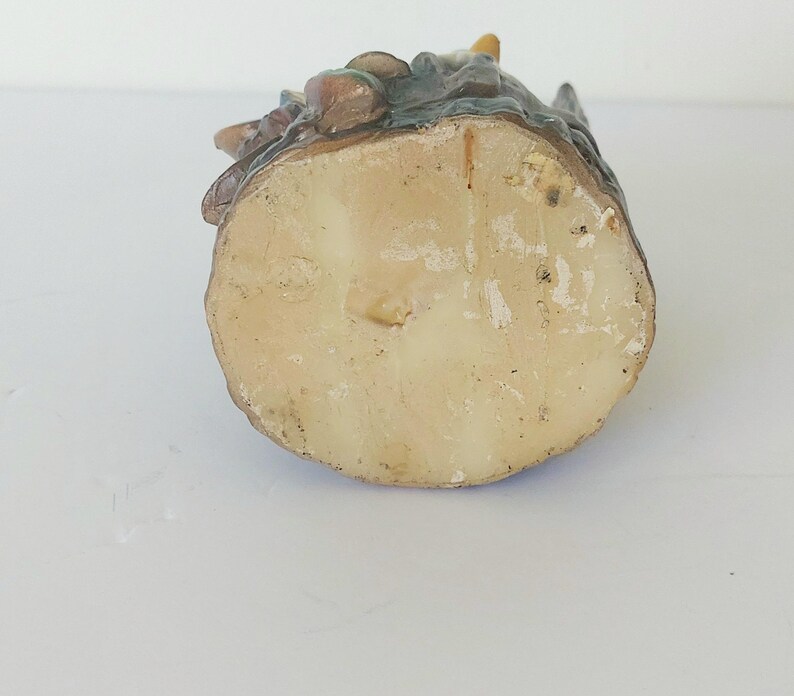 May include: The bottom of a ceramic figurine of a gnome sitting on a tree stump. The figurine is painted in shades of brown, green, and blue. The bottom of the figurine is white and has a rough texture.