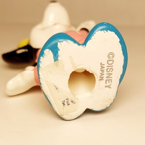 May include: A white ceramic figurine of a cartoon dog wearing a pink dress and a blue hat. The figurine is standing on its hind legs and has a hole in the bottom. The bottom of the figurine is marked with the text "&copy;DISNEY JAPAN".