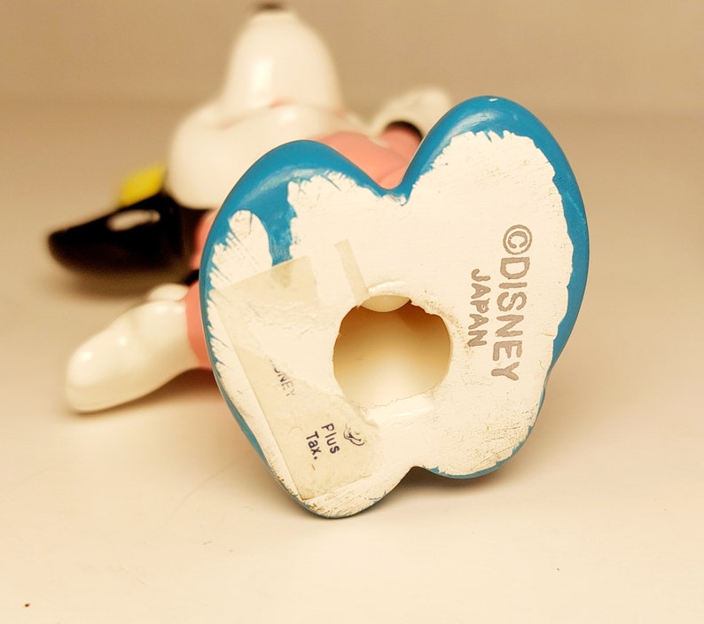 May include: A white ceramic figurine of a cartoon dog wearing a pink dress and a blue hat. The figurine is standing on its hind legs and has a hole in the bottom. The bottom of the figurine is marked with the text "&copy;DISNEY JAPAN".